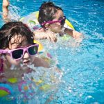 Two children with sunglasses float and splash in a bright blue pool